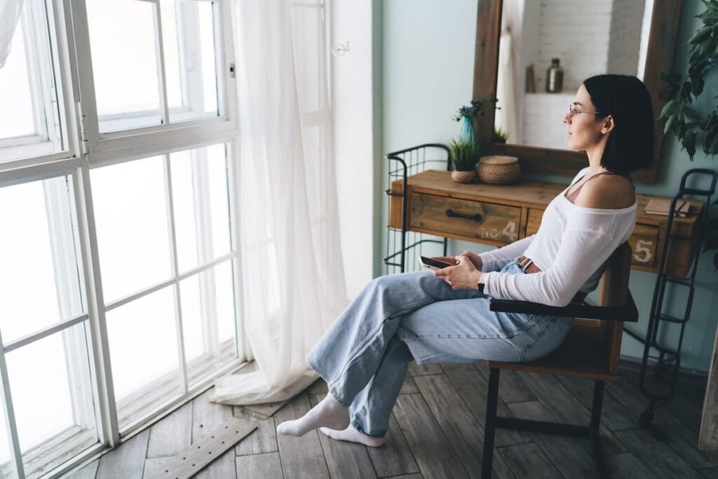 Woman sitting by a window, using her phone and reflecting quietly with a moment of pause during sociopolitical stress.